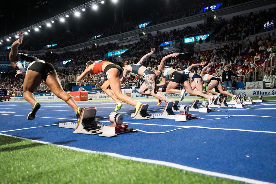 Start der 60-Meter-Hürdenläuferinnen auf einer blauen Laufbahn, Zuschauer im Hintergrund.