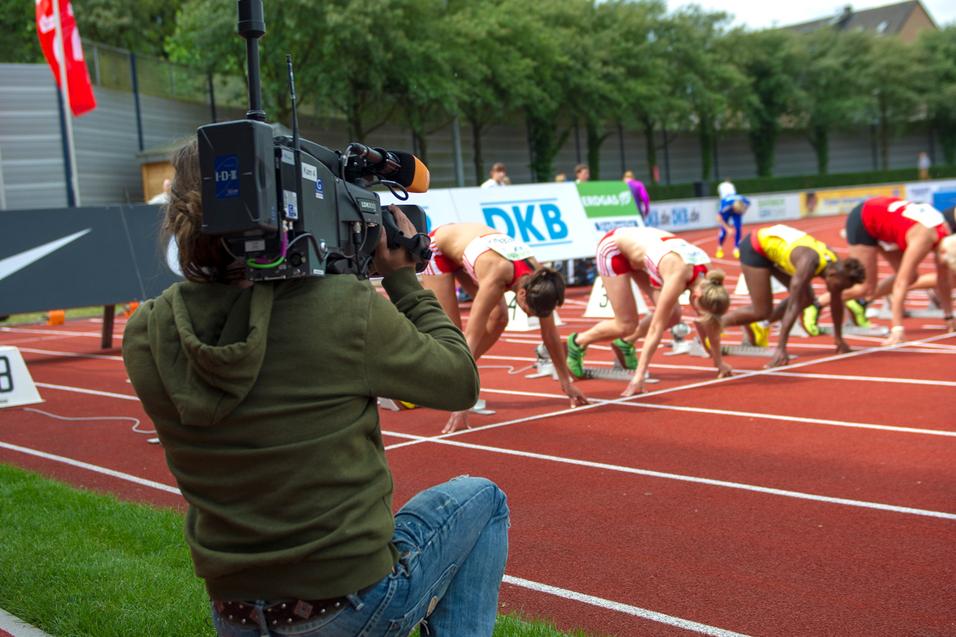Kameramann filmt sprintende Athletinnen an der Startlinie auf einer leuchtend roten Tartanbahn.